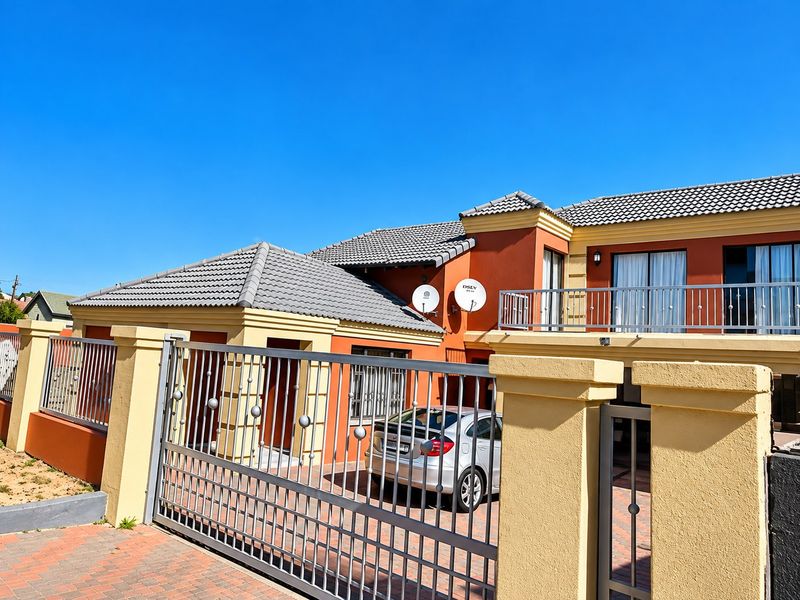 Front elevation of a two-story house featuring a paved driveway, secure metal gate, tiled roof, and an upper-level balcony with metal railings.