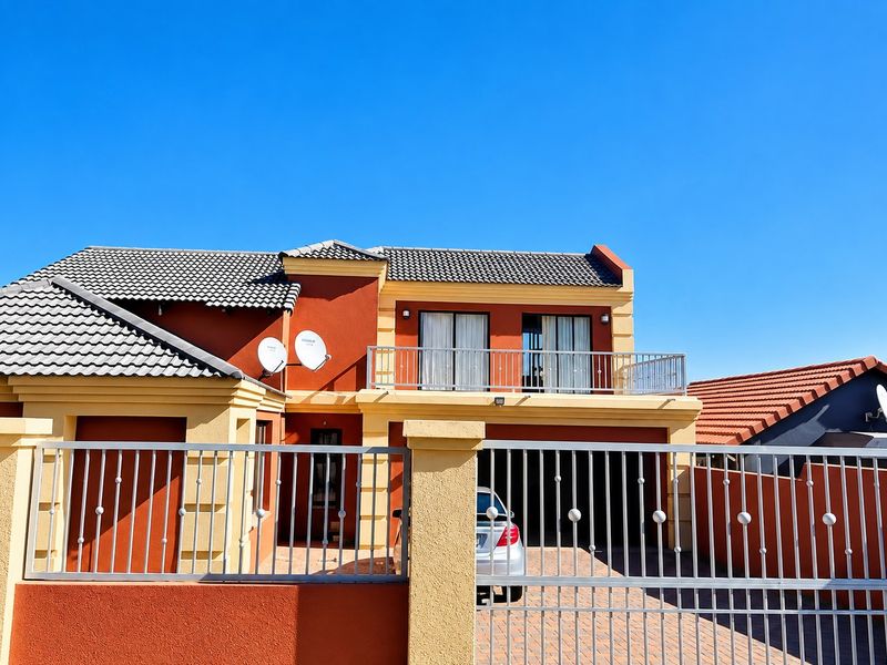 Front elevation of a two-story house featuring a tiled roof, balcony with metal railing, and a paved driveway with secure perimeter fencing.