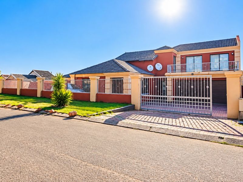 Front elevation of a two-story residential house featuring a paved driveway, secure metal gate, boundary wall, and an upper-level balcony.