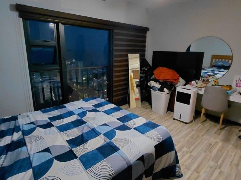A bedroom featuring laminate flooring, a ceiling fan, and a sliding glass door with roller blinds, offering a view of the city at night.