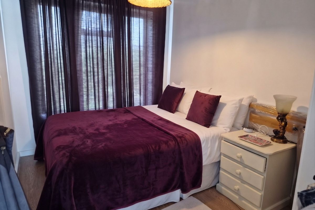 A cozy bedroom featuring laminate flooring and a central pendant light fixture, with large windows dressed in dark curtains.