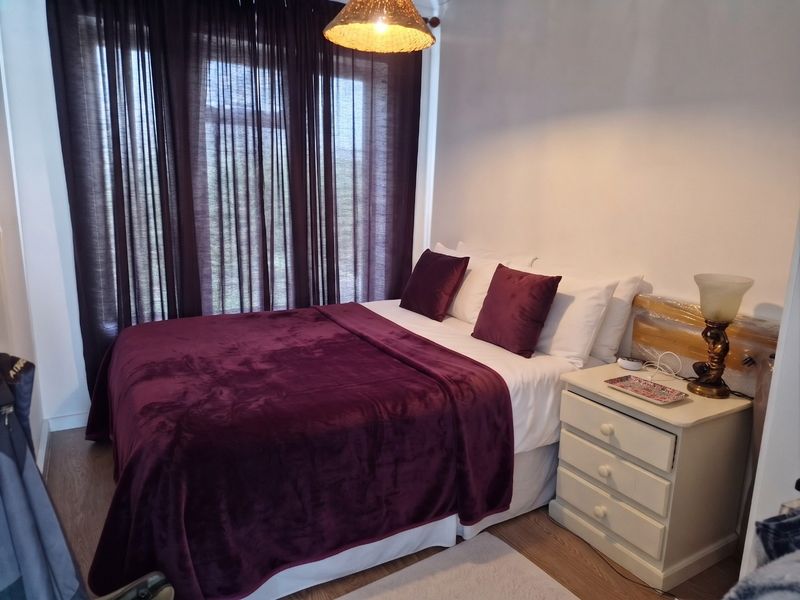 A cozy bedroom featuring laminate flooring and a central pendant light fixture, with large windows dressed in dark curtains.