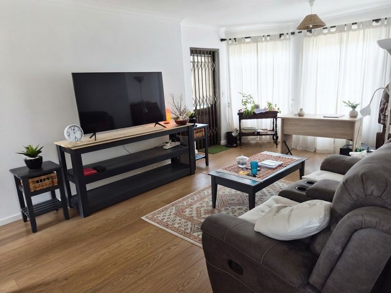 A well-lit living room featuring light-toned laminate flooring, neutral walls, and a security gate leading to an outdoor area.