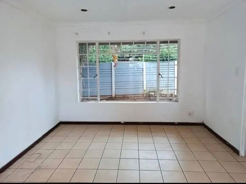 A bright, empty living room featuring neutral tiled flooring, recessed lighting, and a large window with security bars overlooking the exterior.
