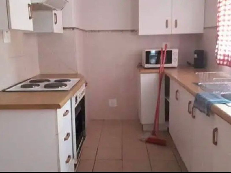 A functional galley-style kitchen featuring white cabinetry, laminate countertops, an electric hob, built-in oven, and a stainless steel sink with tiled flooring.