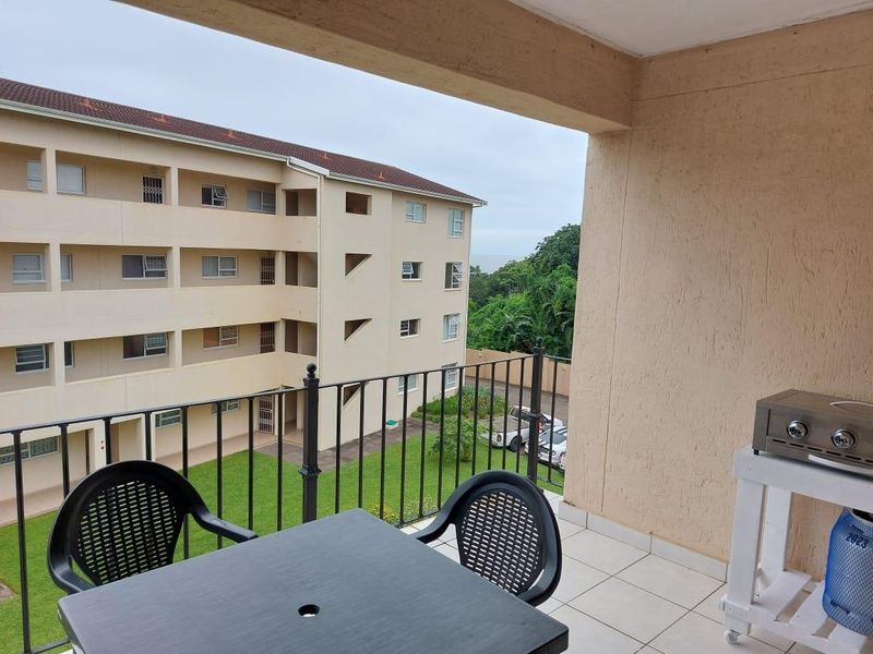 A covered balcony featuring tiled flooring and a metal balustrade, overlooking the apartment complex grounds and surrounding greenery.