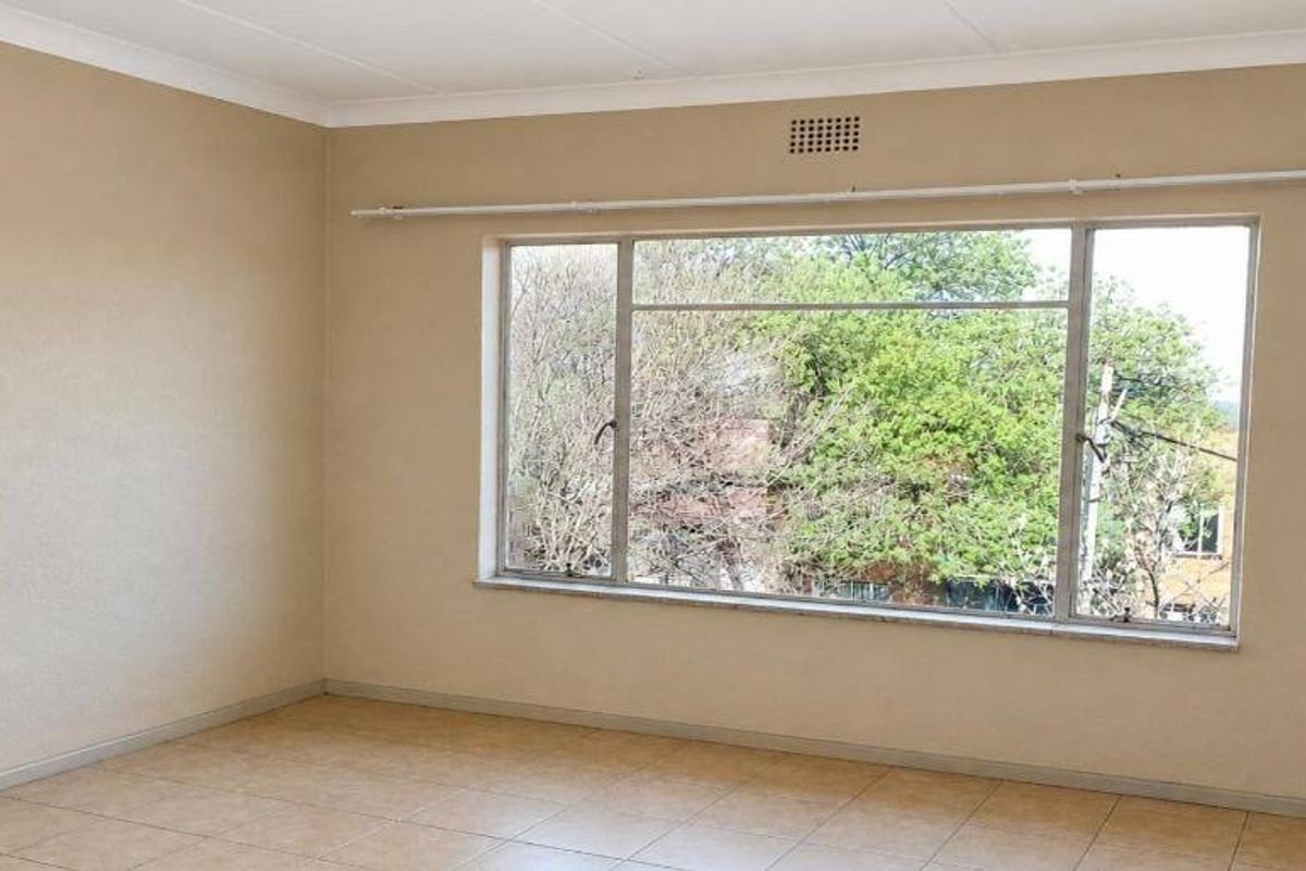 A spacious, empty bedroom featuring neutral wall tones, tiled flooring, and a large window with a curtain rail, offering a bright and versatile living space.