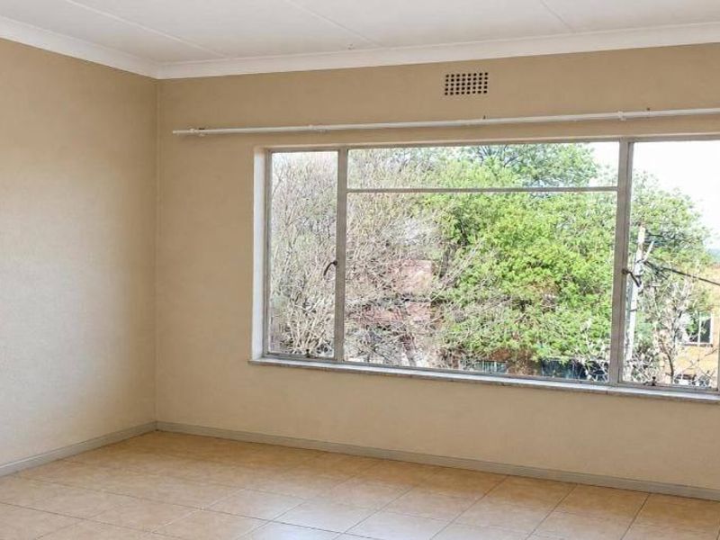 A spacious, empty bedroom featuring neutral wall tones, tiled flooring, and a large window with a curtain rail, offering a bright and versatile living space.