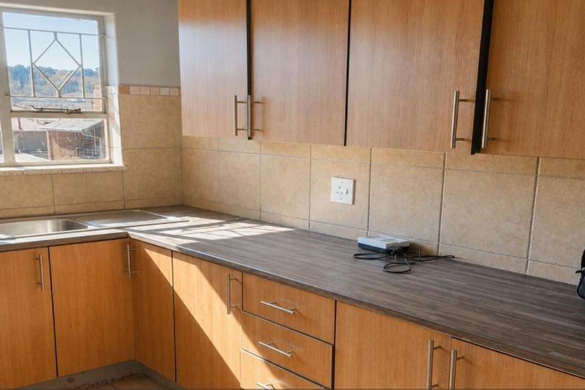 A functional kitchen featuring wood-finish cabinetry, laminate countertops, a stainless steel double sink, and tiled backsplash with natural light from the window.