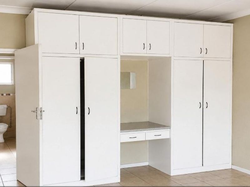 A clean bedroom featuring extensive white built-in cupboards with an integrated vanity desk, tiled flooring, and direct access to an en-suite bathroom.