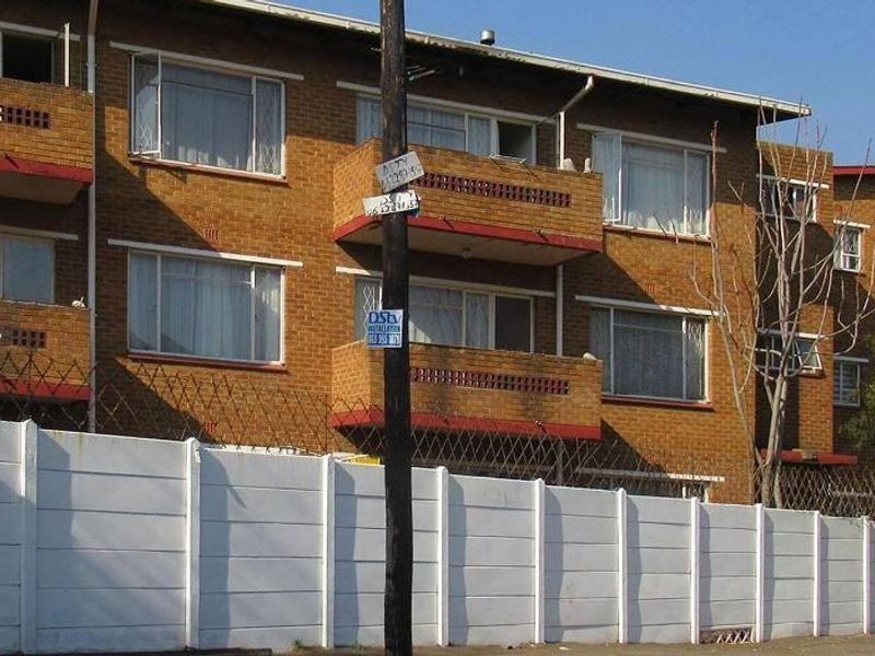Exterior view of a multi-story apartment building featuring face brick construction, private balconies, and a secure perimeter wall with razor wire.