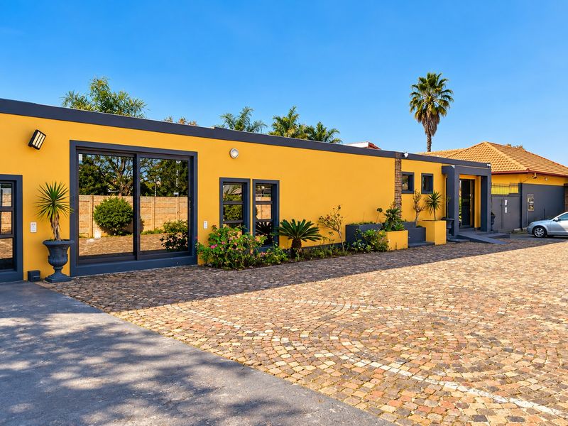 A well-maintained commercial building exterior featuring a vibrant yellow facade, modern dark-framed sliding glass doors, and a spacious cobblestone paved parking area.