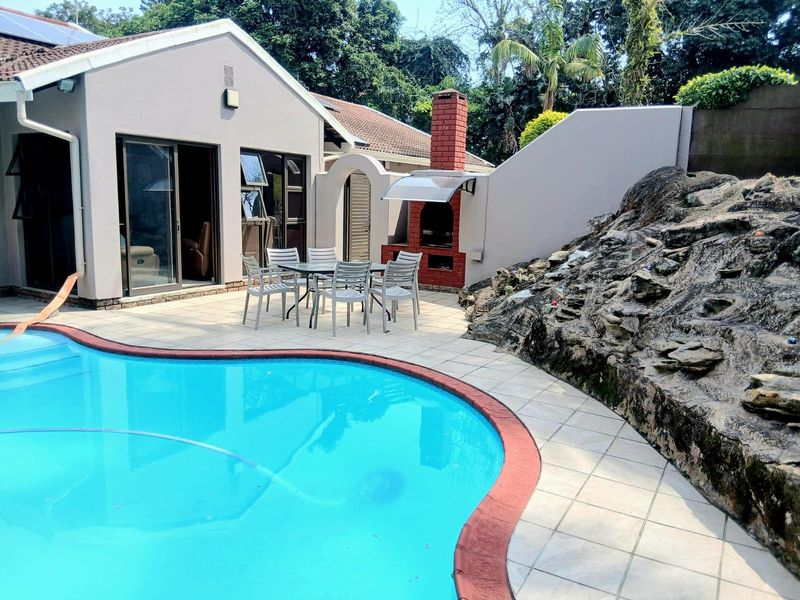 Outdoor entertainment area featuring a swimming pool, tiled patio, and a built-in brick braai, set against a natural rock feature.