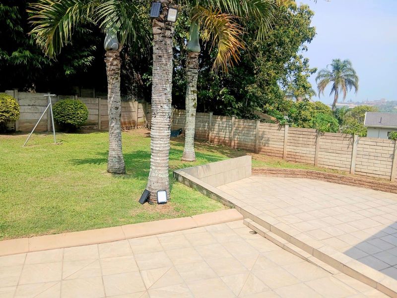 A well-maintained garden featuring a tiled patio area, retaining wall, and established palm trees, enclosed by a secure precast concrete boundary wall.