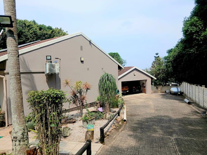 Rear view of the property featuring a paved driveway, a garage, and a landscaped garden area with established trees and shrubs.