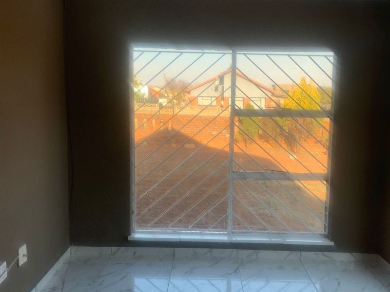 A bright, empty living room featuring modern marble-look floor tiles, recessed downlights, and a large window fitted with security burglar bars.