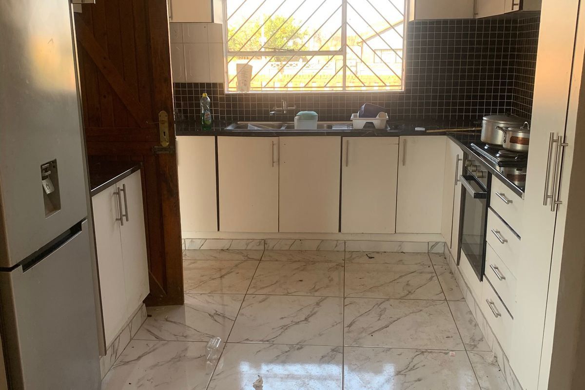 A functional kitchen featuring white cabinetry, black tiled backsplash, marble-effect floor tiles, and integrated appliances, illuminated by natural light through a window with security bars.