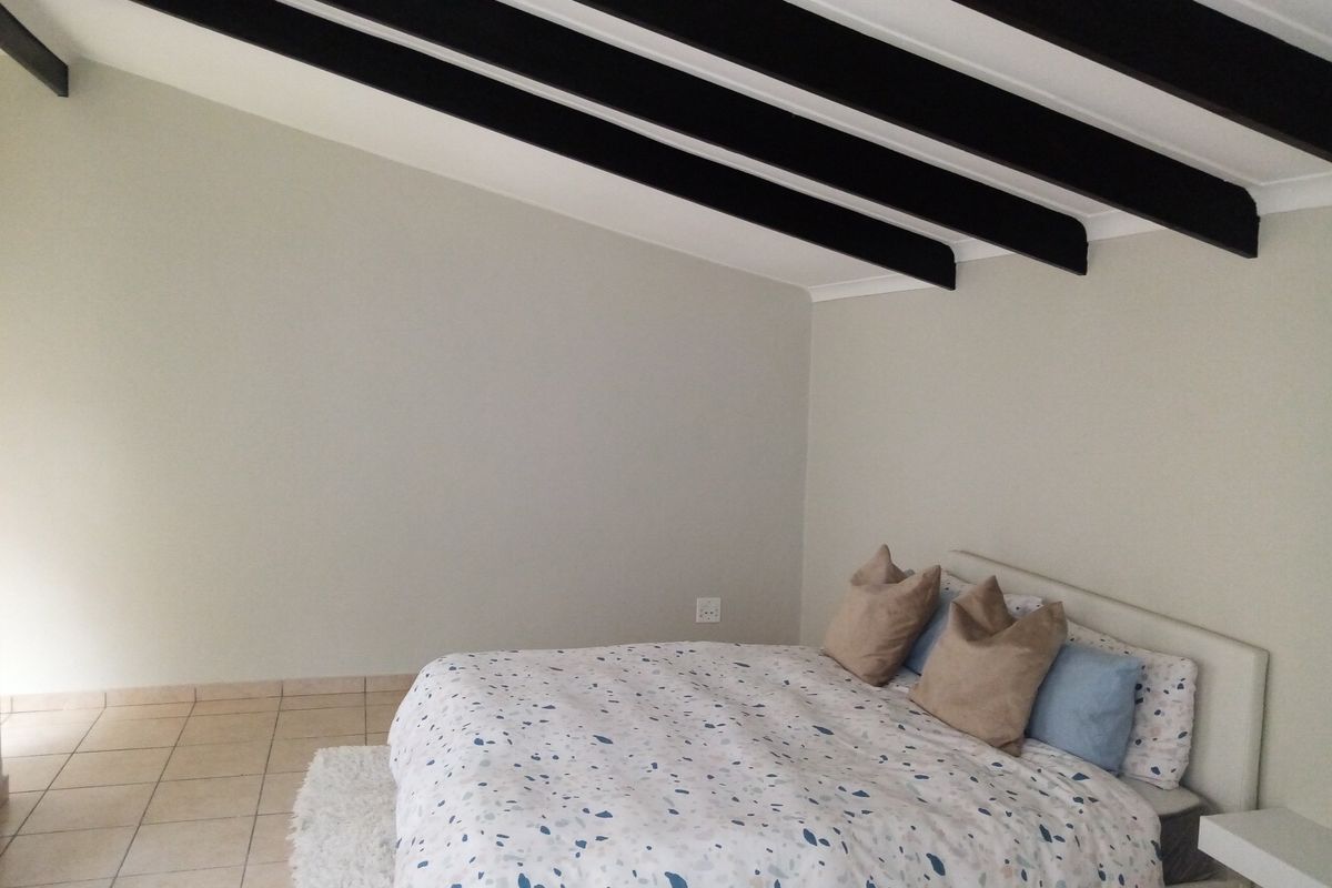 A bedroom featuring a raked ceiling with exposed dark timber beams and neutral tiled flooring.
