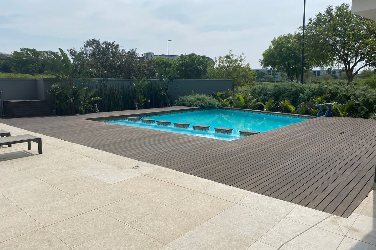 Outdoor pool area featuring a swimming pool surrounded by timber decking and stone paving, enclosed by a boundary wall.