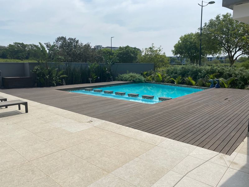 Outdoor pool area featuring a swimming pool surrounded by timber decking and stone paving, enclosed by a boundary wall.