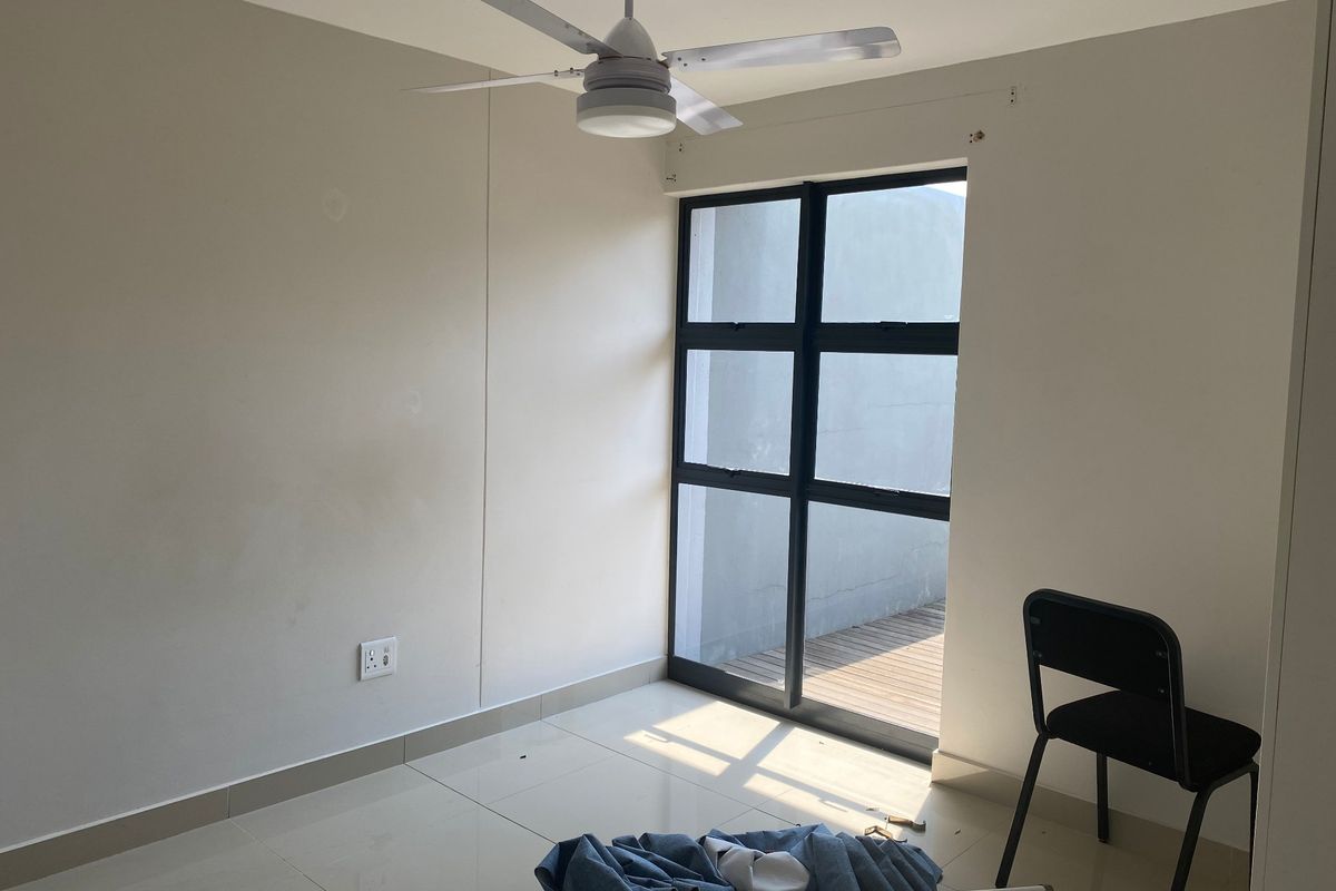 A bright, empty bedroom featuring tiled flooring, a ceiling fan, and a large black aluminium window with access to an outdoor area.