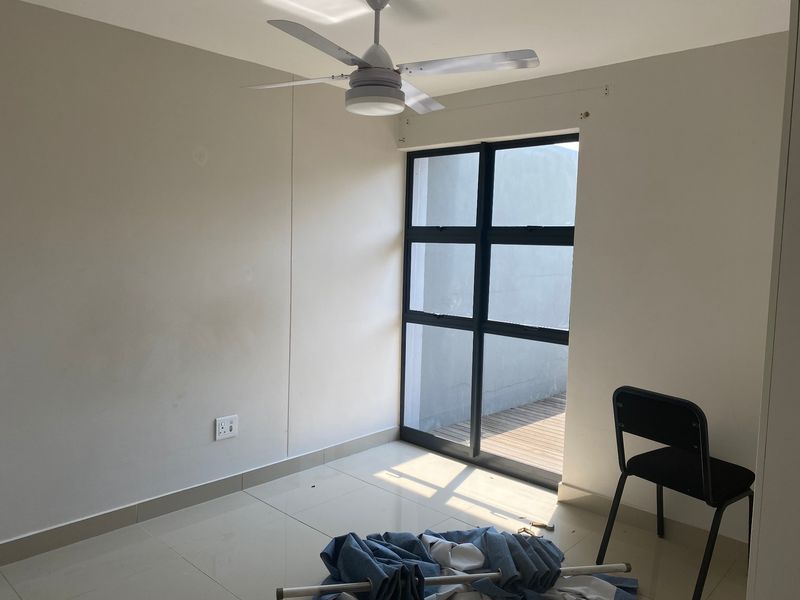 A bright, empty bedroom featuring tiled flooring, a ceiling fan, and a large black aluminium window with access to an outdoor area.
