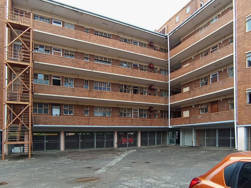 Exterior view of a multi-story brick apartment building featuring a metal fire escape, ground-level security gates, and a paved courtyard area.