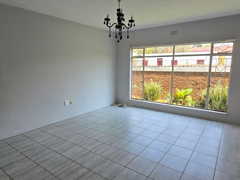 A bright, empty living room featuring neutral tiled flooring, a large window with security bars, and a central ceiling light fixture.
