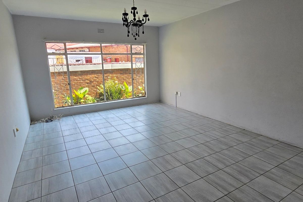 A bright, empty living room featuring neutral tiled flooring, a large window providing natural light, and a central ceiling light fixture.