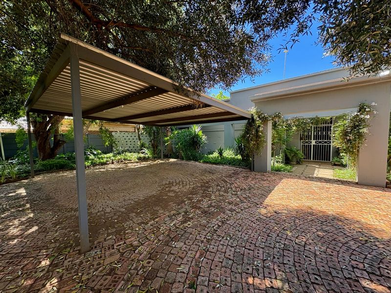 A well-maintained front entrance featuring a paved circular driveway, a functional carport, and a secure gated entryway surrounded by established garden landscaping.