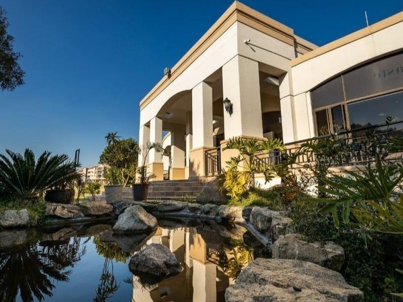A well-maintained front exterior view featuring a decorative stone-lined pond and a covered patio area with architectural pillars.