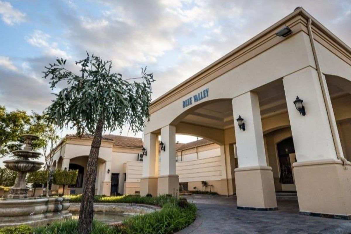 The grand entrance of the Blue Valley estate featuring a covered portico, paved driveway, and a decorative stone fountain in the landscaped front garden.