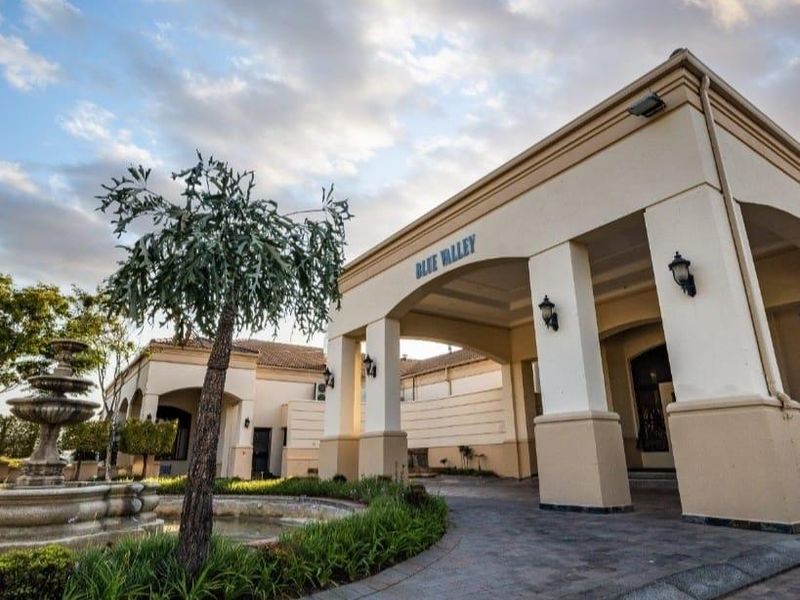 The grand entrance of the Blue Valley estate featuring a covered portico, paved driveway, and a decorative stone fountain in the landscaped front garden.
