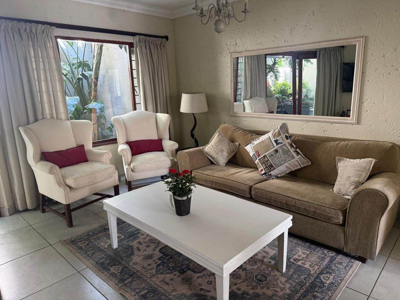 A living room featuring tiled flooring, a large window with garden views, and a wall-mounted mirror, offering a comfortable and functional living space.