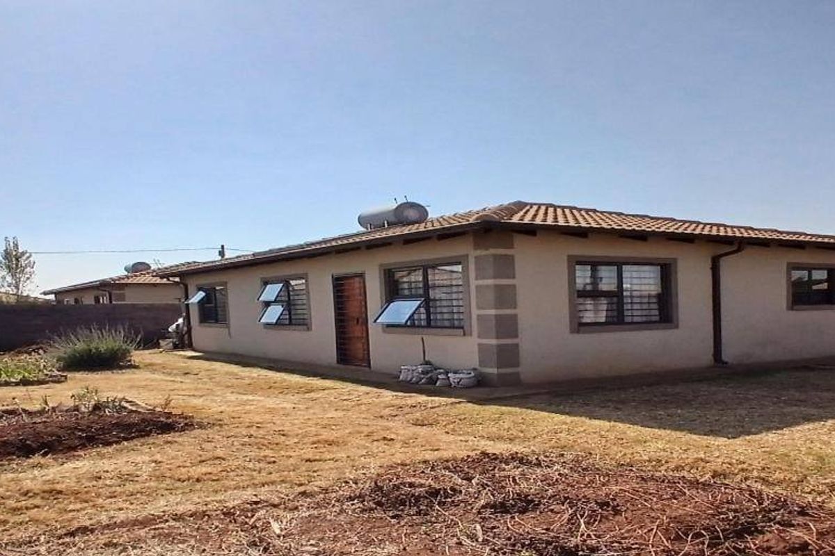 Rear view of a single-story residential house featuring a tiled roof, solar water heater, and a surrounding perimeter wall with a garden area.