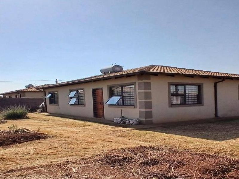 Rear view of a single-story residential house featuring a tiled roof, solar water heater, and a surrounding perimeter wall with a garden area.