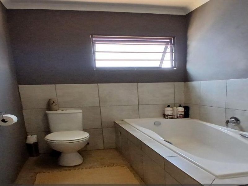 A functional bathroom featuring a built-in bathtub, toilet, and neutral wall tiling with a louvered window for ventilation.