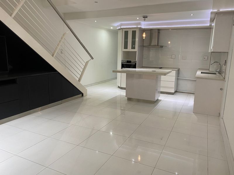 Modern open-plan kitchen and living area featuring sleek white cabinetry, a central island, tiled flooring, and a contemporary staircase with integrated under-stair storage.