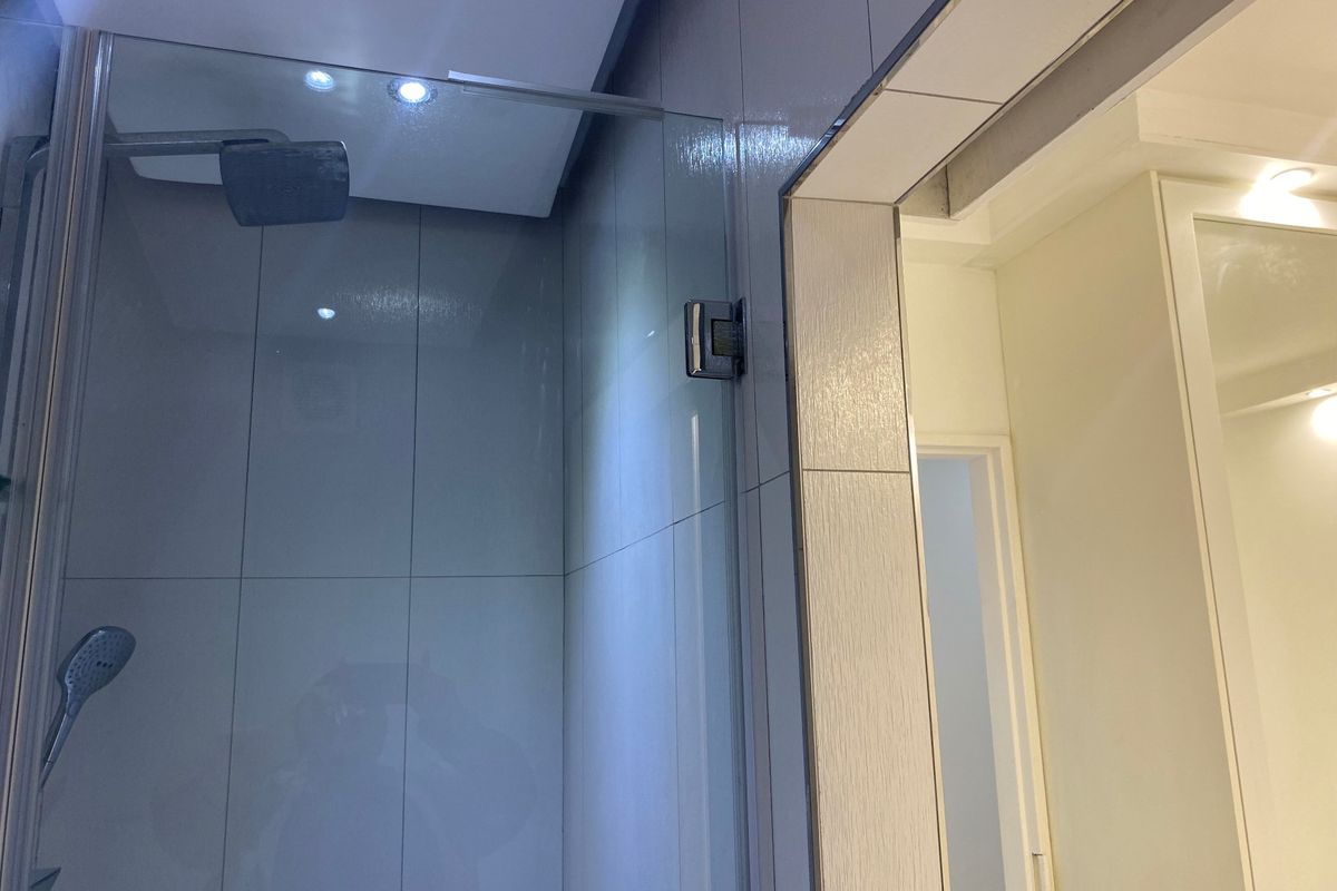 A modern bathroom featuring a glass-enclosed shower with both rain and handheld shower heads, complemented by recessed ceiling lighting and neutral wall tiling.