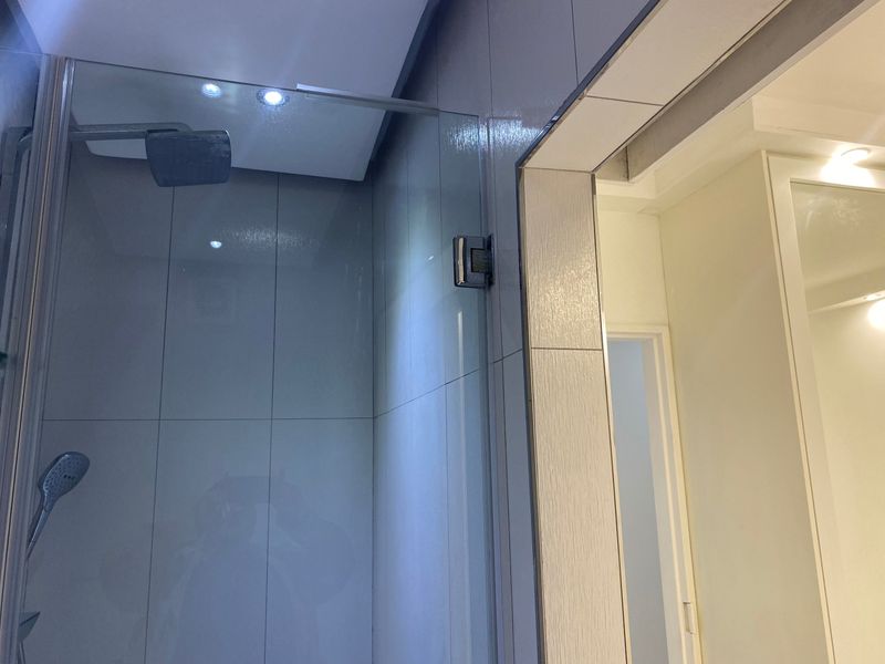 A modern bathroom featuring a glass-enclosed shower with both rain and handheld shower heads, complemented by recessed ceiling lighting and neutral wall tiling.