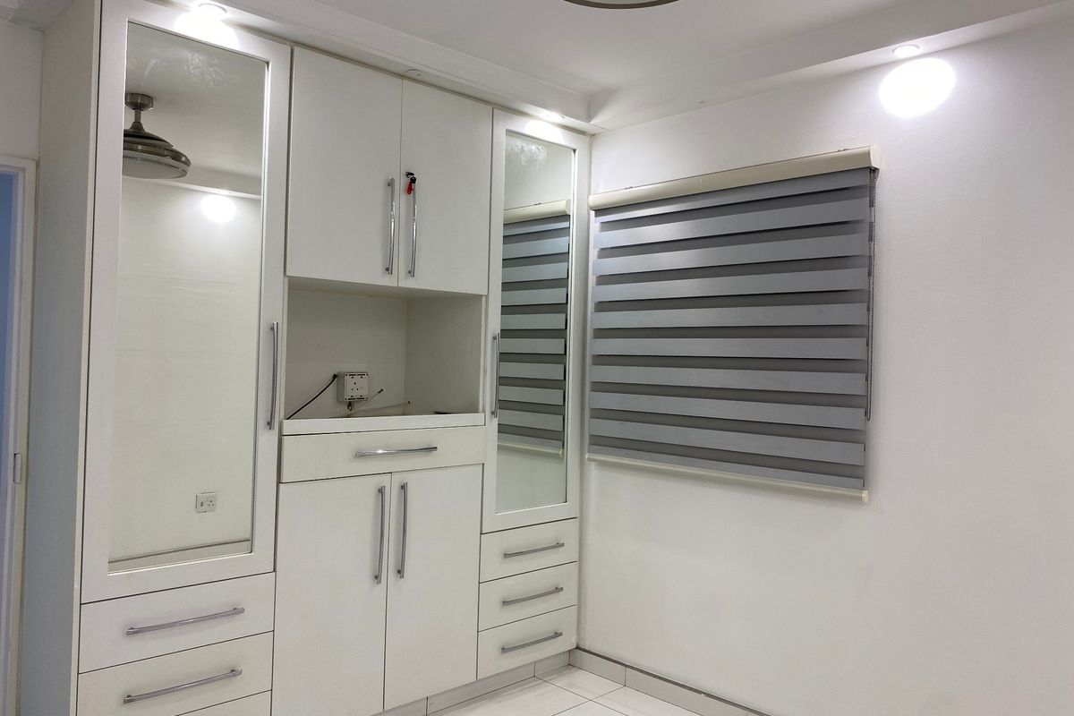 A clean, modern bedroom featuring extensive white built-in cabinetry with integrated mirrors, sleek hardware, and contemporary roller blinds.