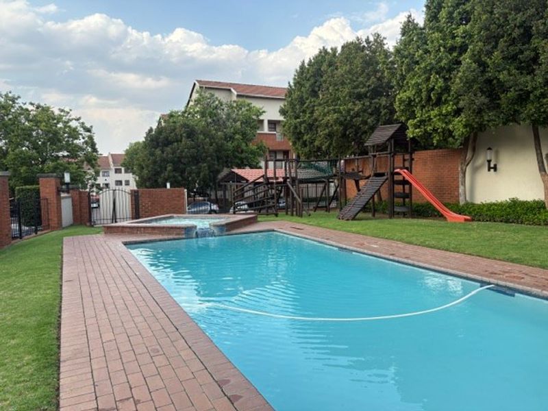 Communal swimming pool area featuring brick paving, a raised water feature, and adjacent landscaped garden space with playground equipment.