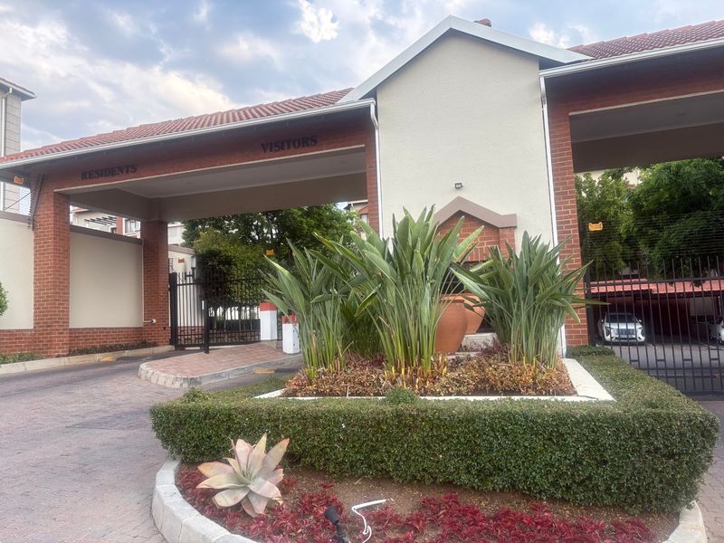 The main entrance to the apartment complex features a secure gated access point with dedicated lanes for residents and visitors, complemented by landscaped gardens.