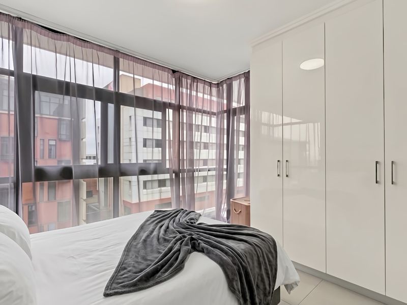 A bright bedroom featuring floor-to-ceiling windows with urban views and sleek, white built-in wardrobes providing ample storage space.