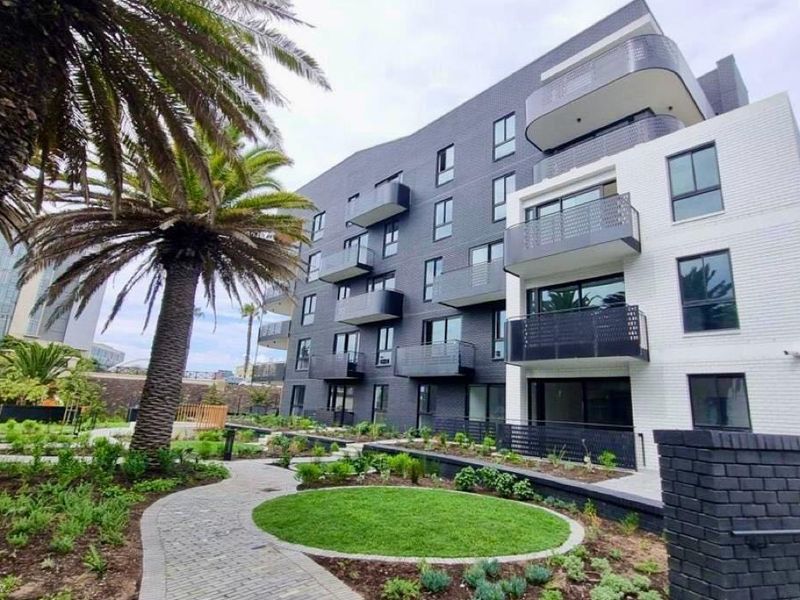 Modern apartment building exterior featuring a landscaped garden, paved pathways, and private balconies with contemporary brick and glass finishes.