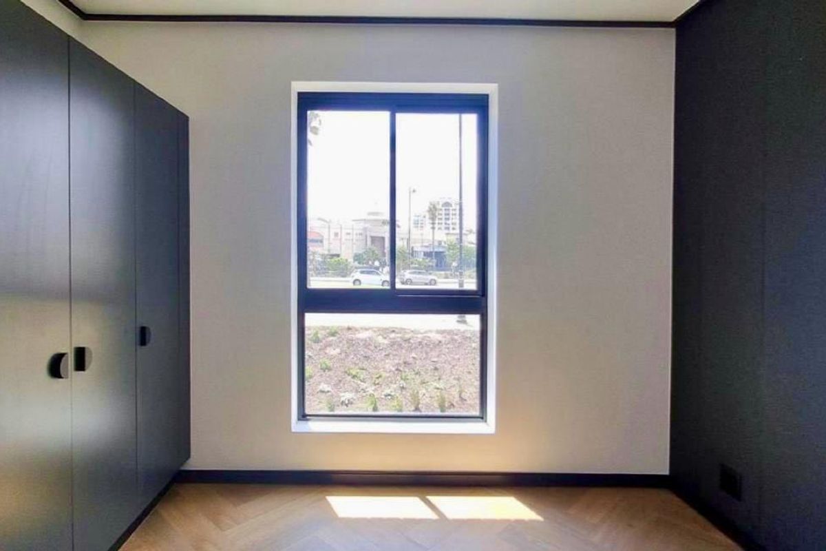 A modern, empty bedroom featuring sleek dark built-in cupboards, herringbone parquet flooring, and a large window providing natural light.