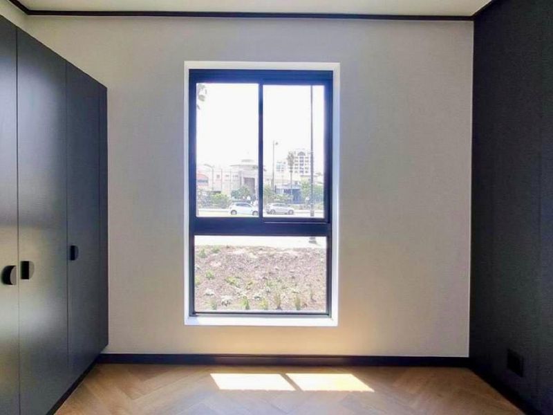 A modern, empty bedroom featuring sleek dark built-in cupboards, herringbone parquet flooring, and a large window providing natural light.