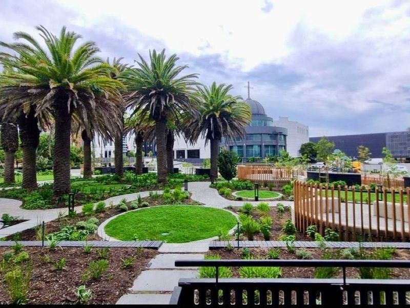 A well-maintained communal garden area featuring mature palm trees, paved walkways, and modern timber-fenced seating zones within an apartment complex.