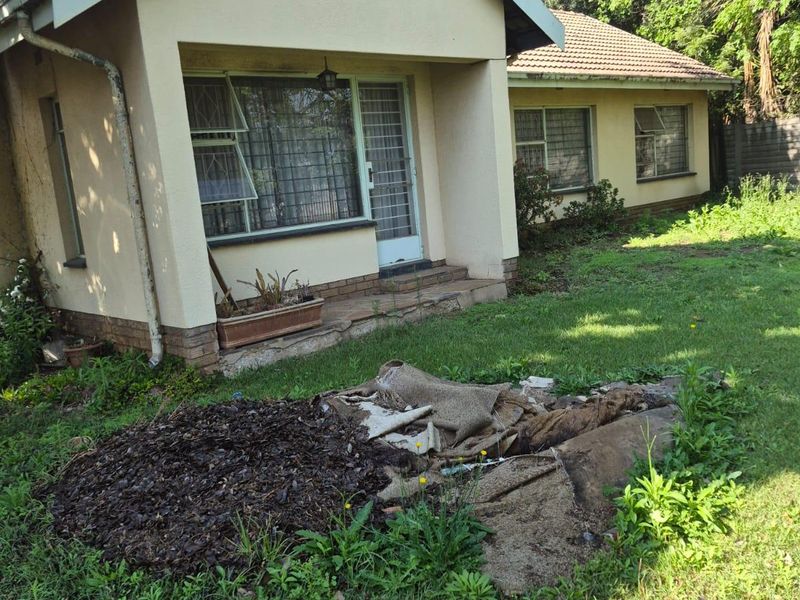 Front elevation of a single-story house featuring a tiled roof, security gates, and burglar bars, set within an overgrown garden requiring maintenance.