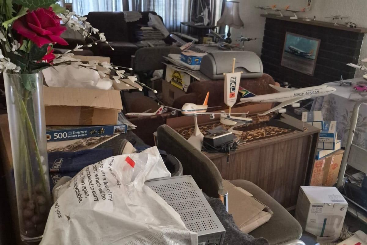 A living room featuring a brick fireplace and tiled flooring, currently cluttered with personal items and requiring significant clearing and renovation.